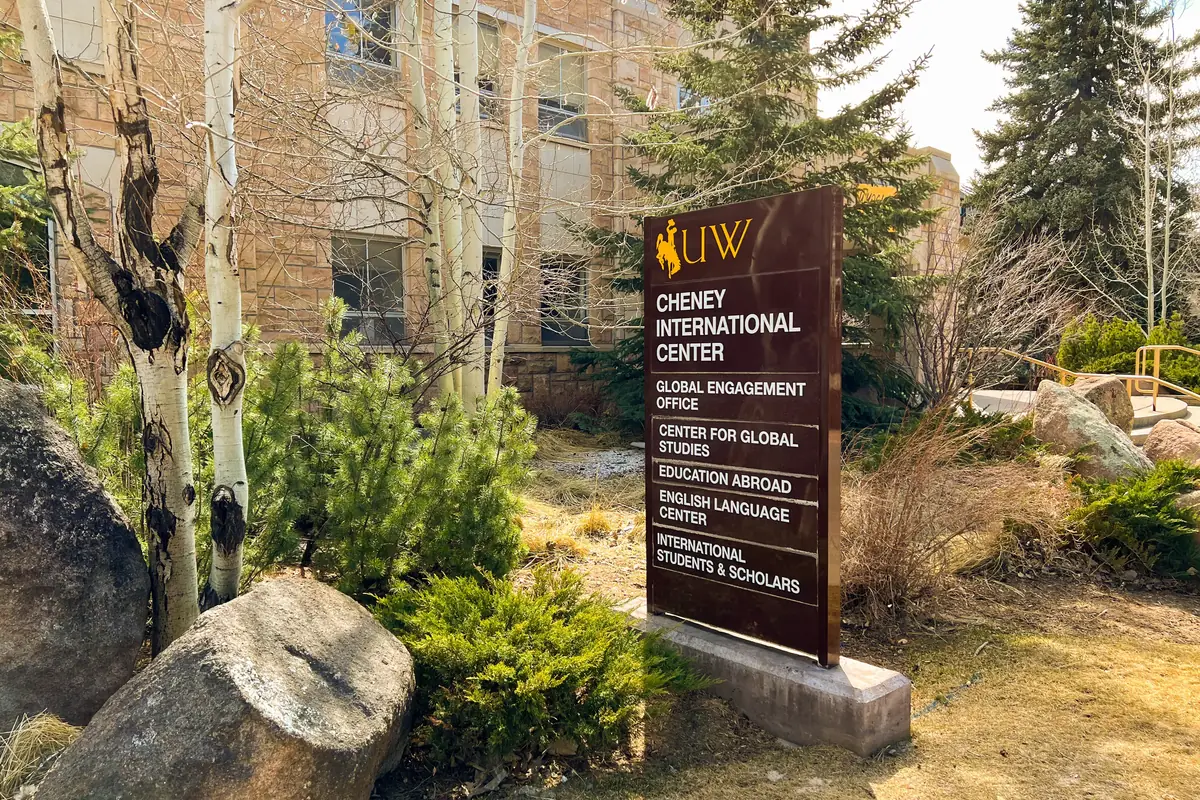 A campus sign for the Cheney International Center at the University of Wyoming, listing offices including International Students and Scholars, among trees and boulders