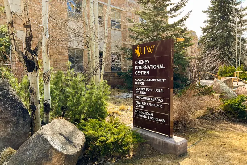 A campus sign for the Cheney International Center at the University of Wyoming, listing offices including International Students and Scholars, among trees and boulders
