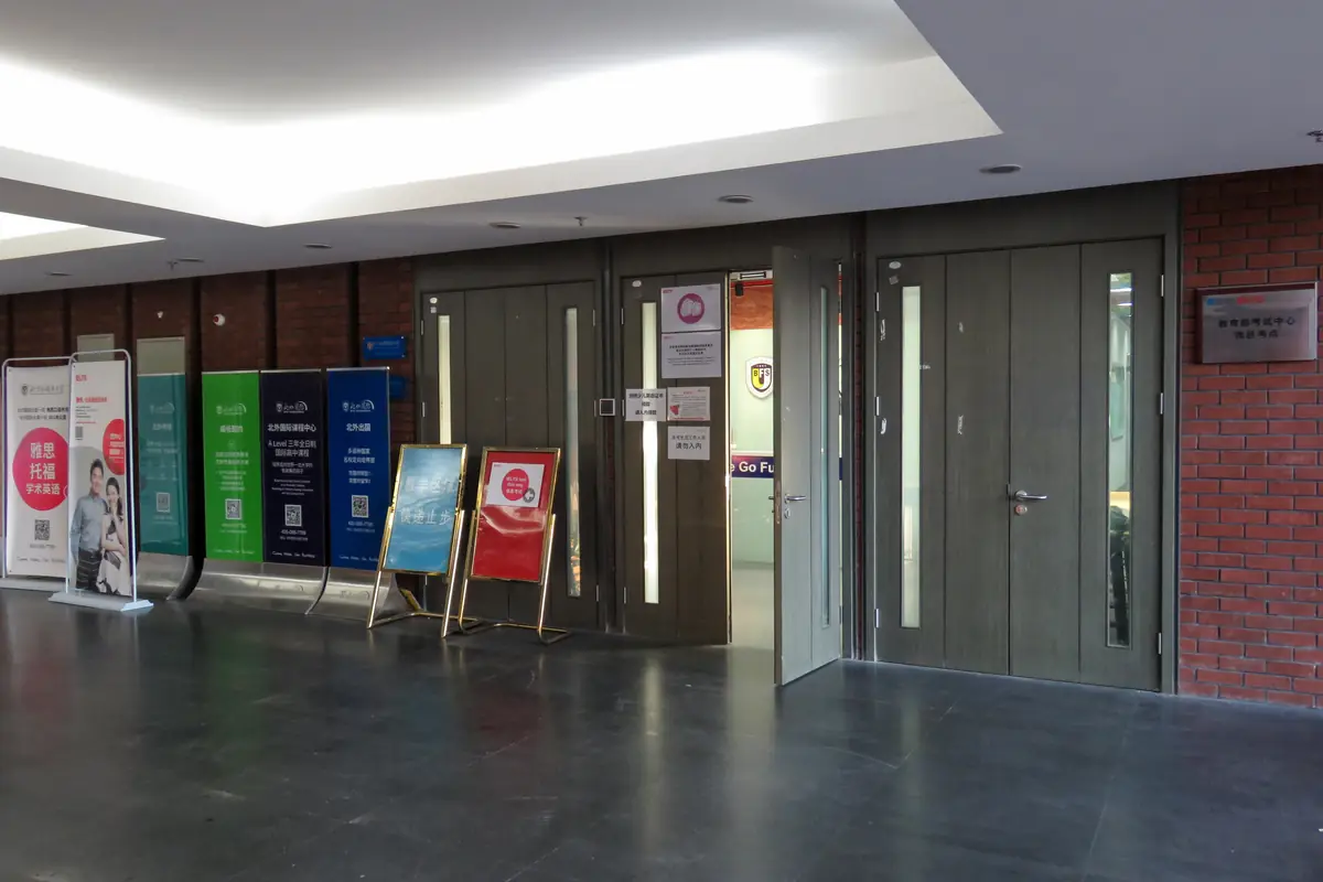 The entrance to an IELTS test centre in a university corridor: open grey doors, notice boards and exam posters along a brick wall
