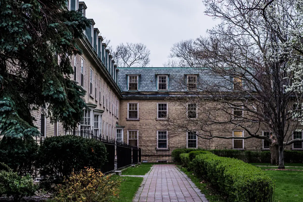 A brick path between clipped hedges leads to a pale brick college building with dormer windows at the University of Toronto