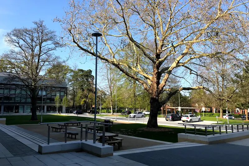 A paved piazza with picnic tables and a large plane tree in front of a modern teaching building on the Oxford Brookes University campus
