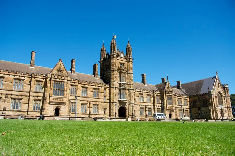 The sandstone Main Quadrangle building of the University of Sydney behind a wide lawn under a blue sky